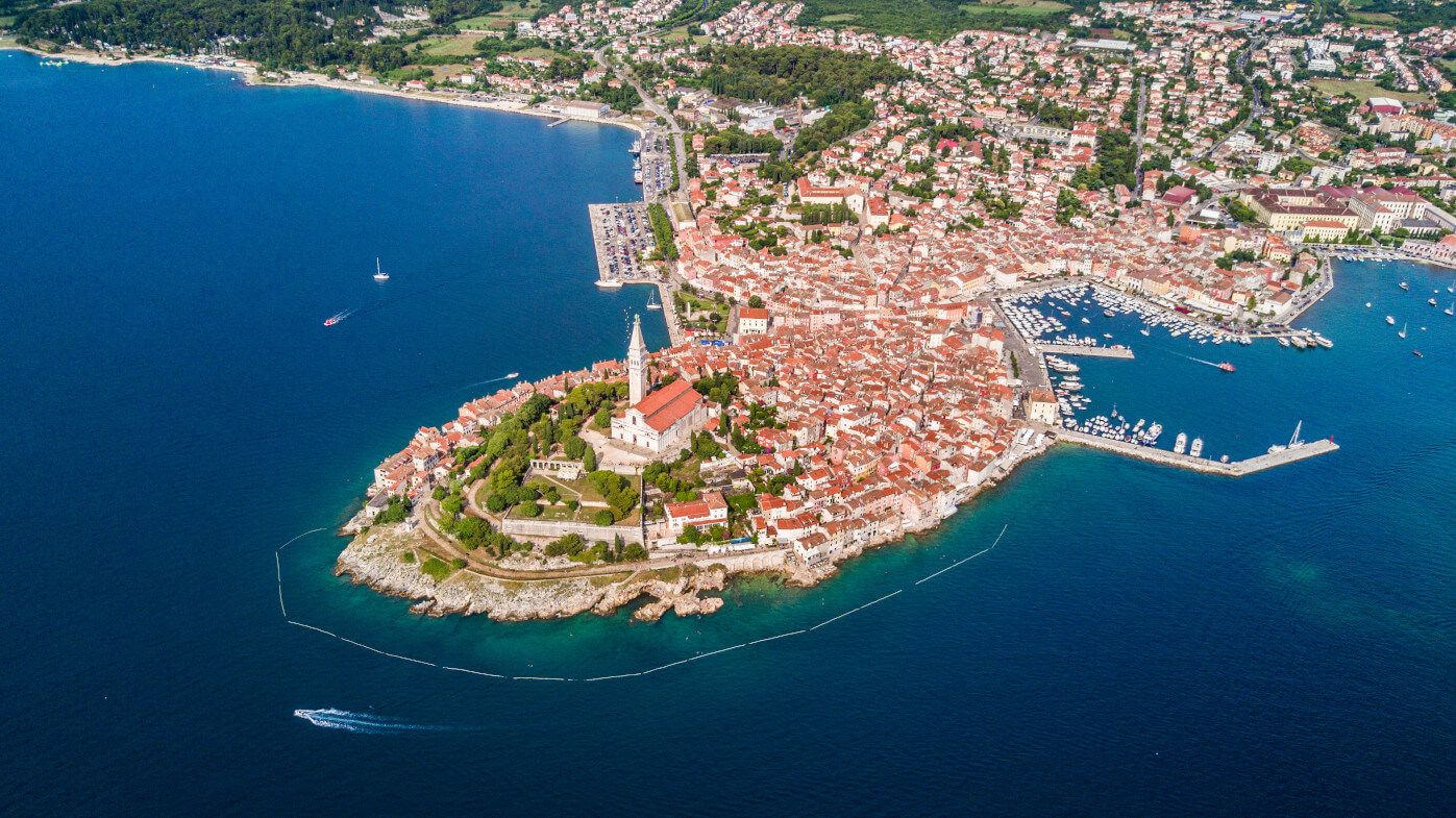 Luftaufnahme einer malerischen Küstenstadt mit historischen Gebäuden, umgeben von blauem Wasser und grünen Hügeln.