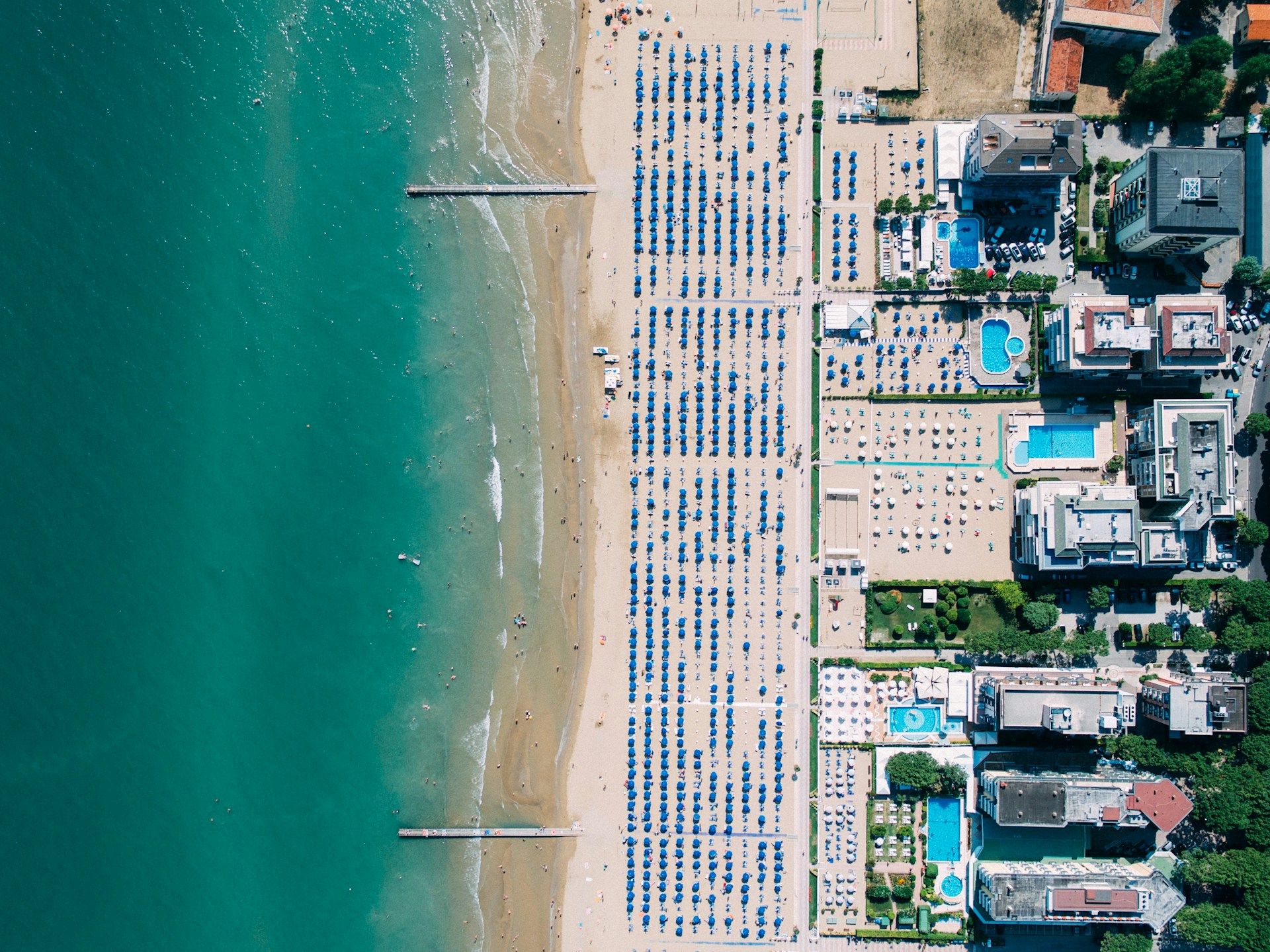 Ein Luftbild zeigt einen Strand mit vielen Sonnenliegen, blauen Schirmen und angrenzenden Hotels sowie Pools.