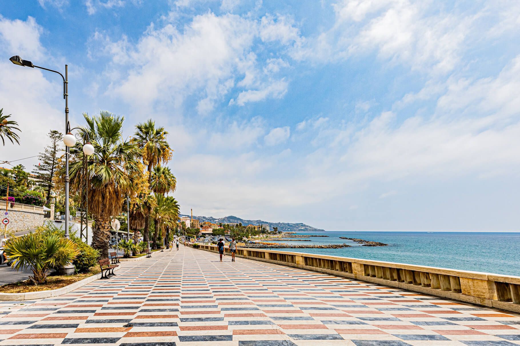 Eine gepflasterte Promenade mit Palmen, Meerblick und bewölktem Himmel, belebte Spaziergänger am Ufer.