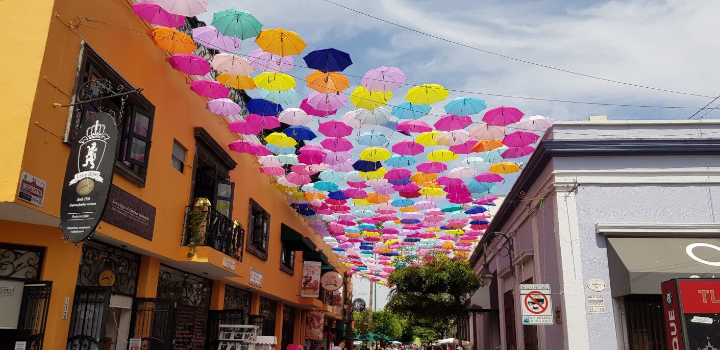 Bunte Regenschirme hängen über einer Straße, gesäumt von farbenfrohen Gebäuden und einem blauen Himmel.