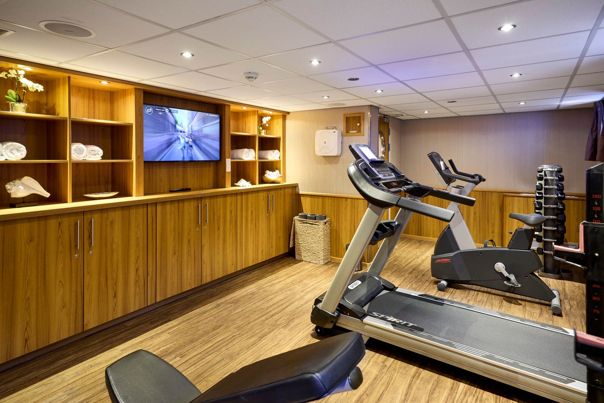 Ein modernes Fitnessstudio mit Laufband, Ergometer und hellem Holzregal. Ein TV-Bildschirm ist an der Wand montiert.