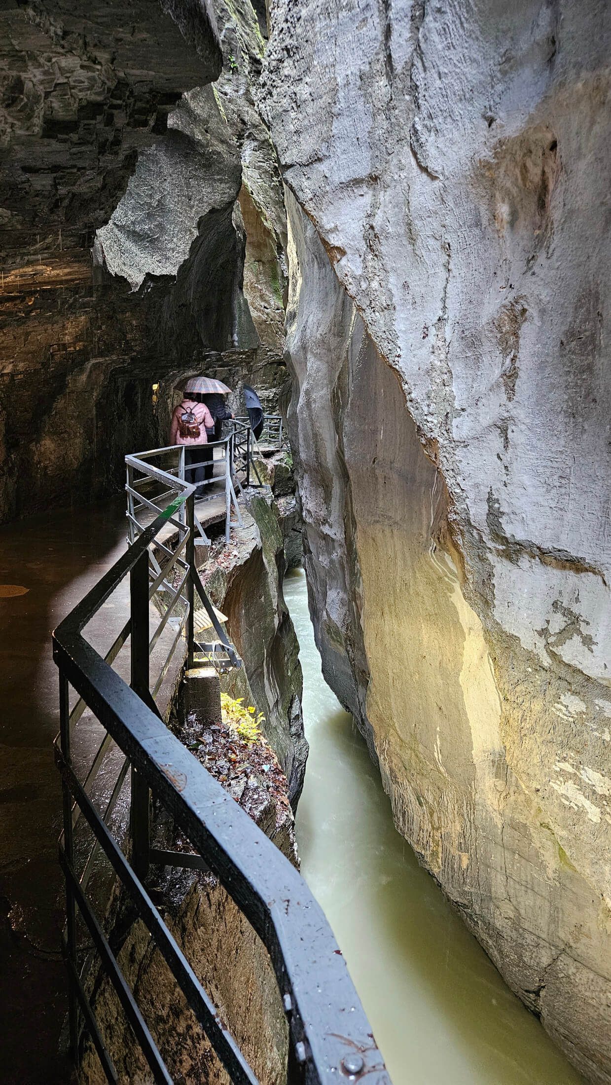 Eine Person mit Regenschirm geht auf einem Weg zwischen steilen Felsen neben einem fließenden Wasserlauf.