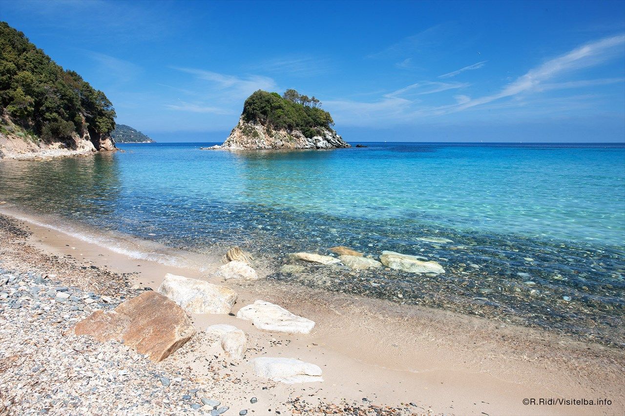 Ein ruhiger Strand auf Elba mit klarem, blauem Wasser und kleinen Inseln im Hintergrund unter einem strahlend blauen Himmel.