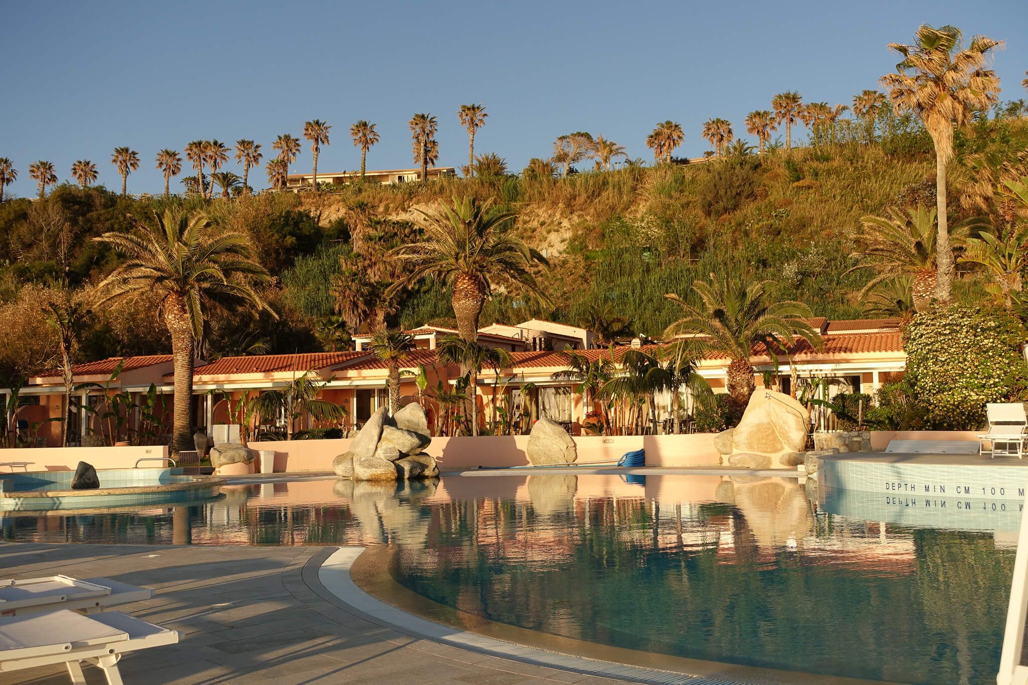Ein Pool mit Palmen und Felsen, umgeben von Häusern und grüner Landschaft, unter einem klaren blauen Himmel.