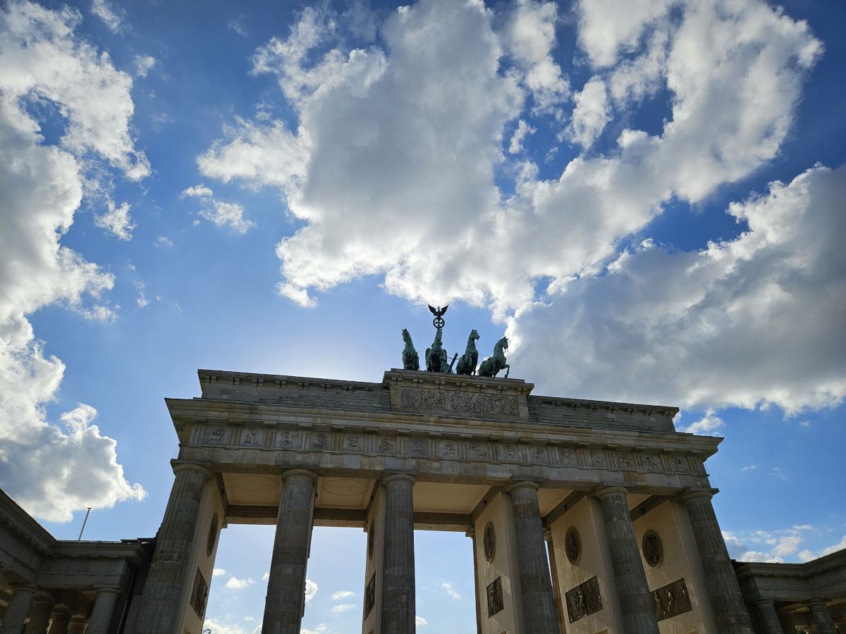 Das Brandenburger Tor vor dramatisch bewölktem Himmel, mit einer Statue auf dem Dach und Sonnenstrahlen im Hintergrund.