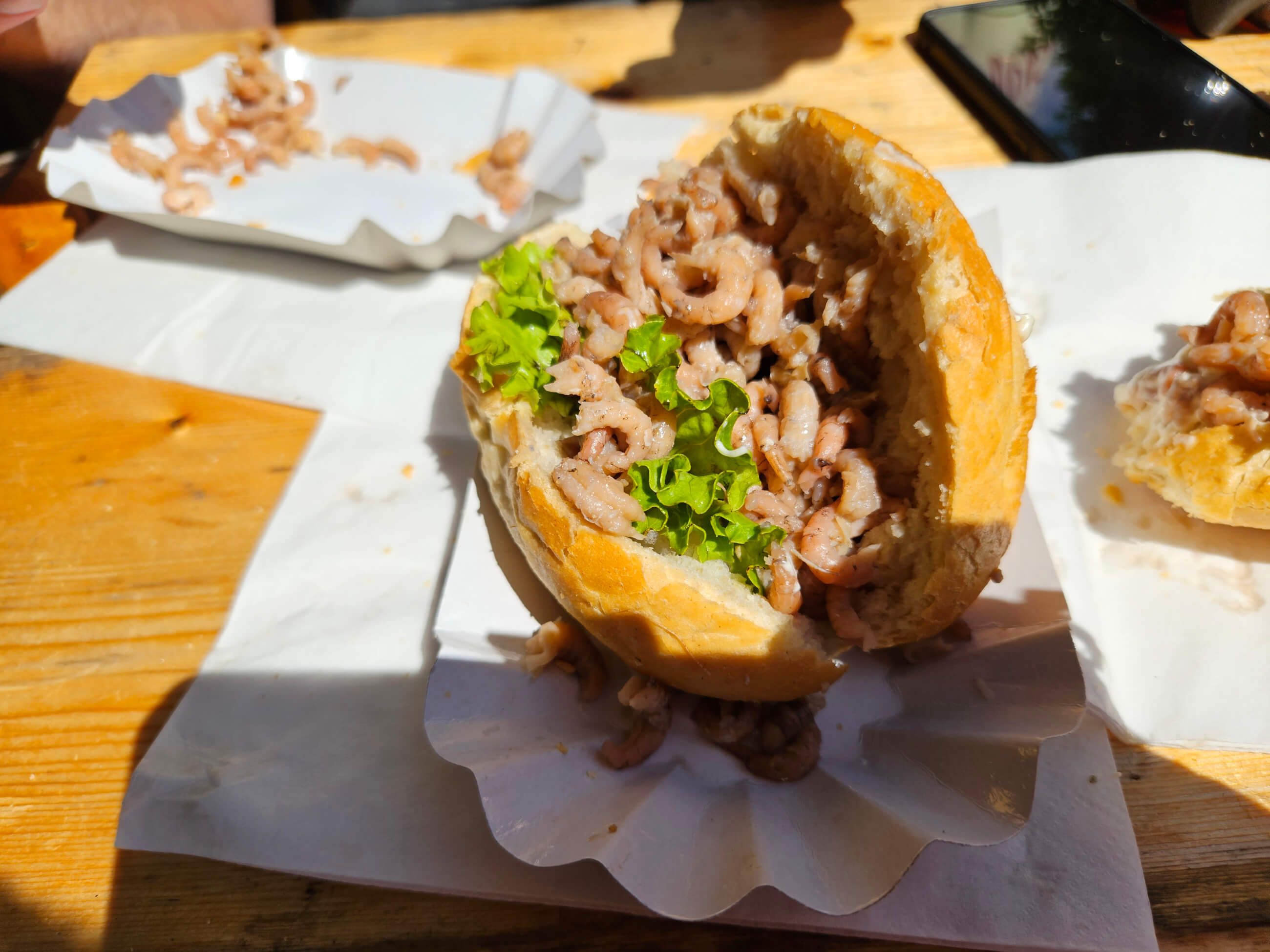 Ein Brötchen gefüllt mit kleinen Garnelen und frischem Salat, auf einem Holztisch serviert.