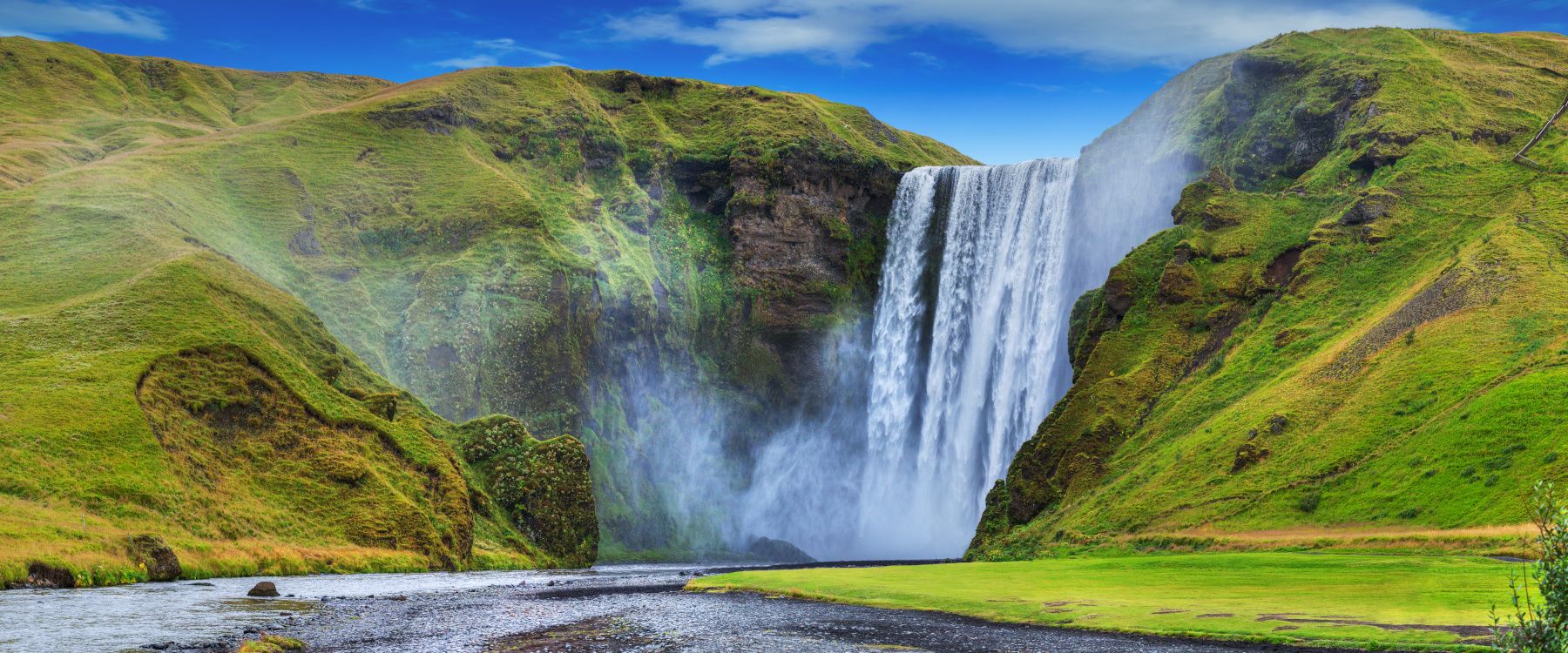 Ein atemberaubender Wasserfall stürzt in eine grüne Landschaft mit Hügeln unter einem klaren blauen Himmel.