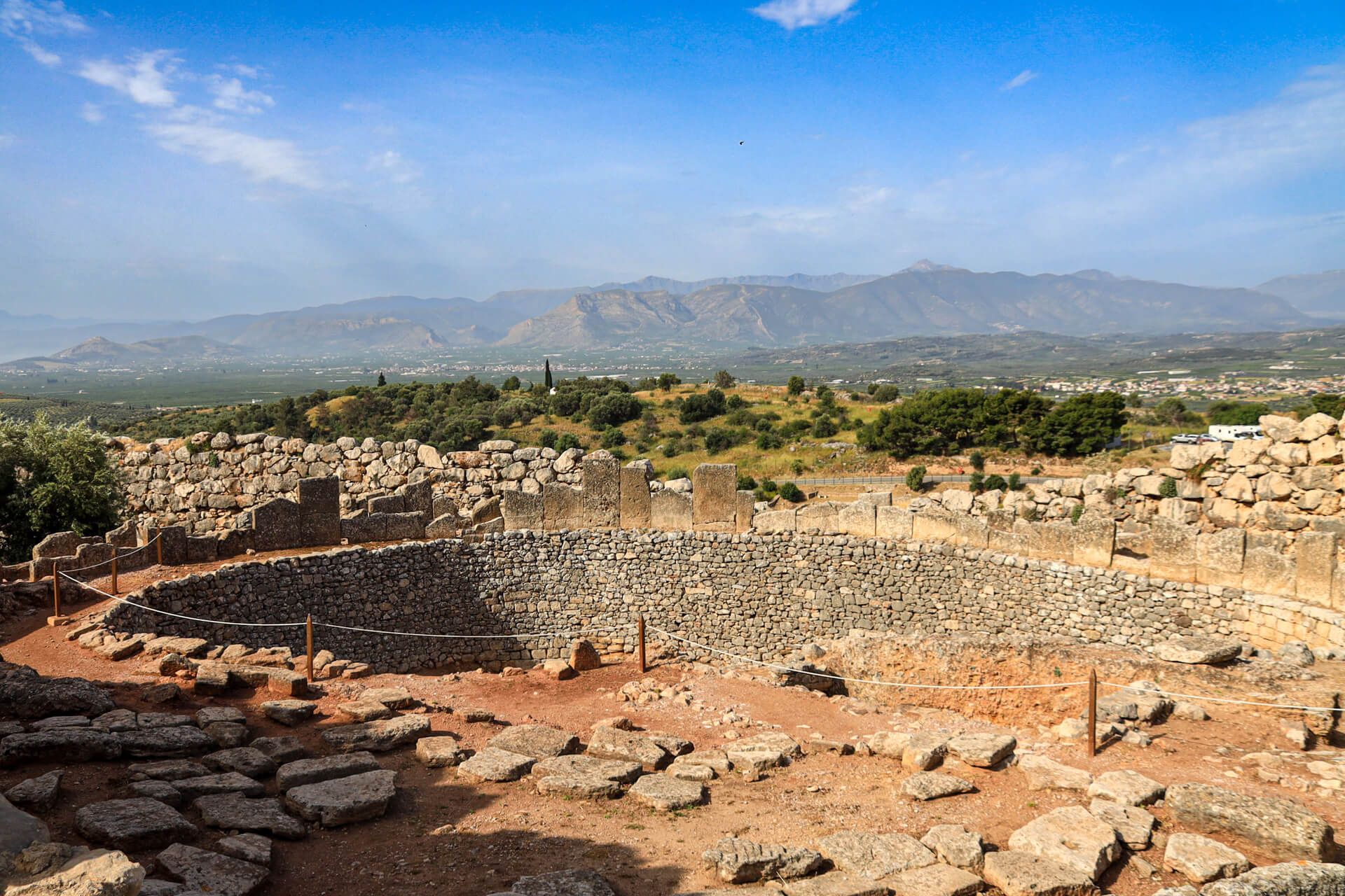 Eine antike Ruine mit runden Mauern, umgeben von Bergen und Grünlandschaft unter blauem Himmel.