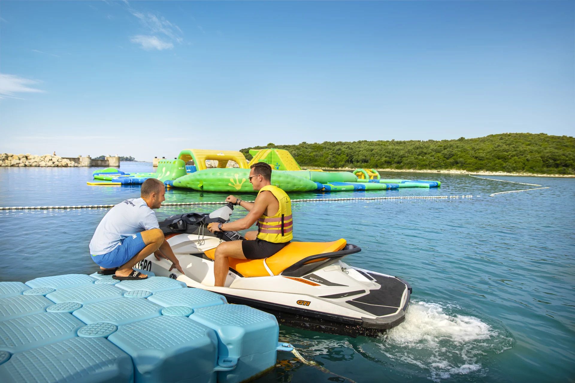 Zwei Männer auf einem Jetski, im Hintergrund bunte Wasserattraktionen und eine klare blaue Lagune.