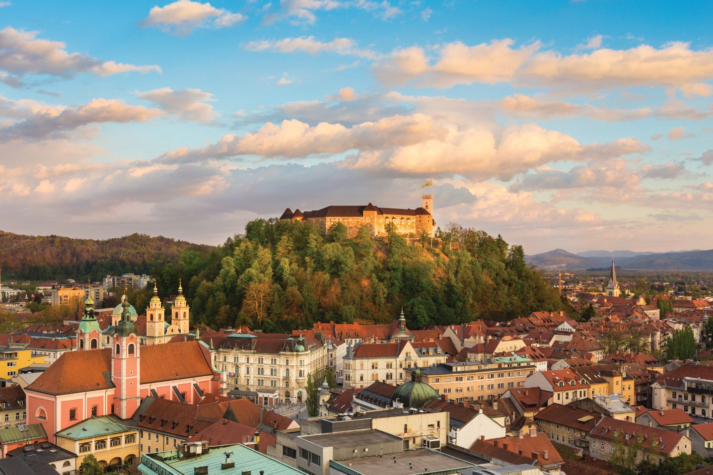 Blick auf die Stadt mit der beeindruckenden Burg auf dem Hügel, umgeben von schönen, grünen Bäumen und leicht bewölktem Himmel.