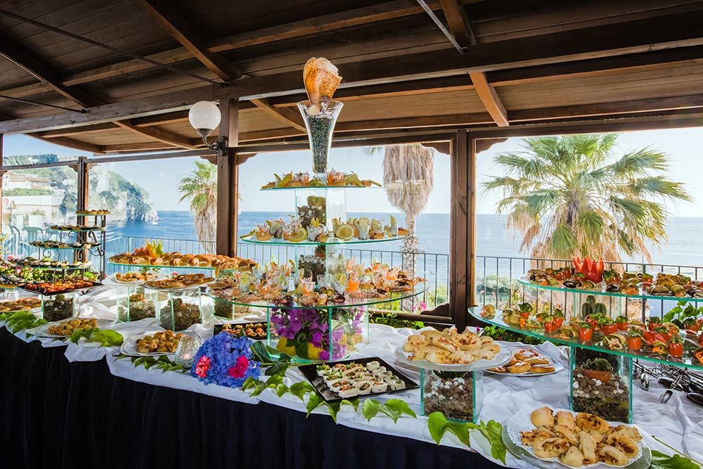 Ein reich gedeckter Tisch mit einer Auswahl an Köstlichkeiten, Blick auf das Meer und Palmen im Hintergrund.