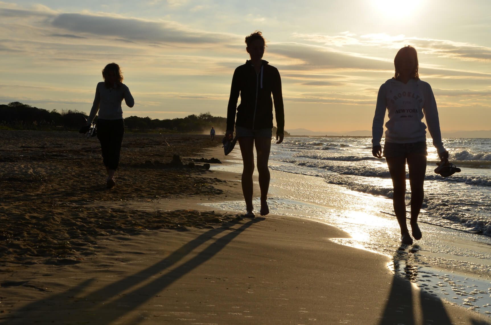 Drei Personen gehen barfuß am Strand entlang, während die Sonne im Hintergrund untergeht.