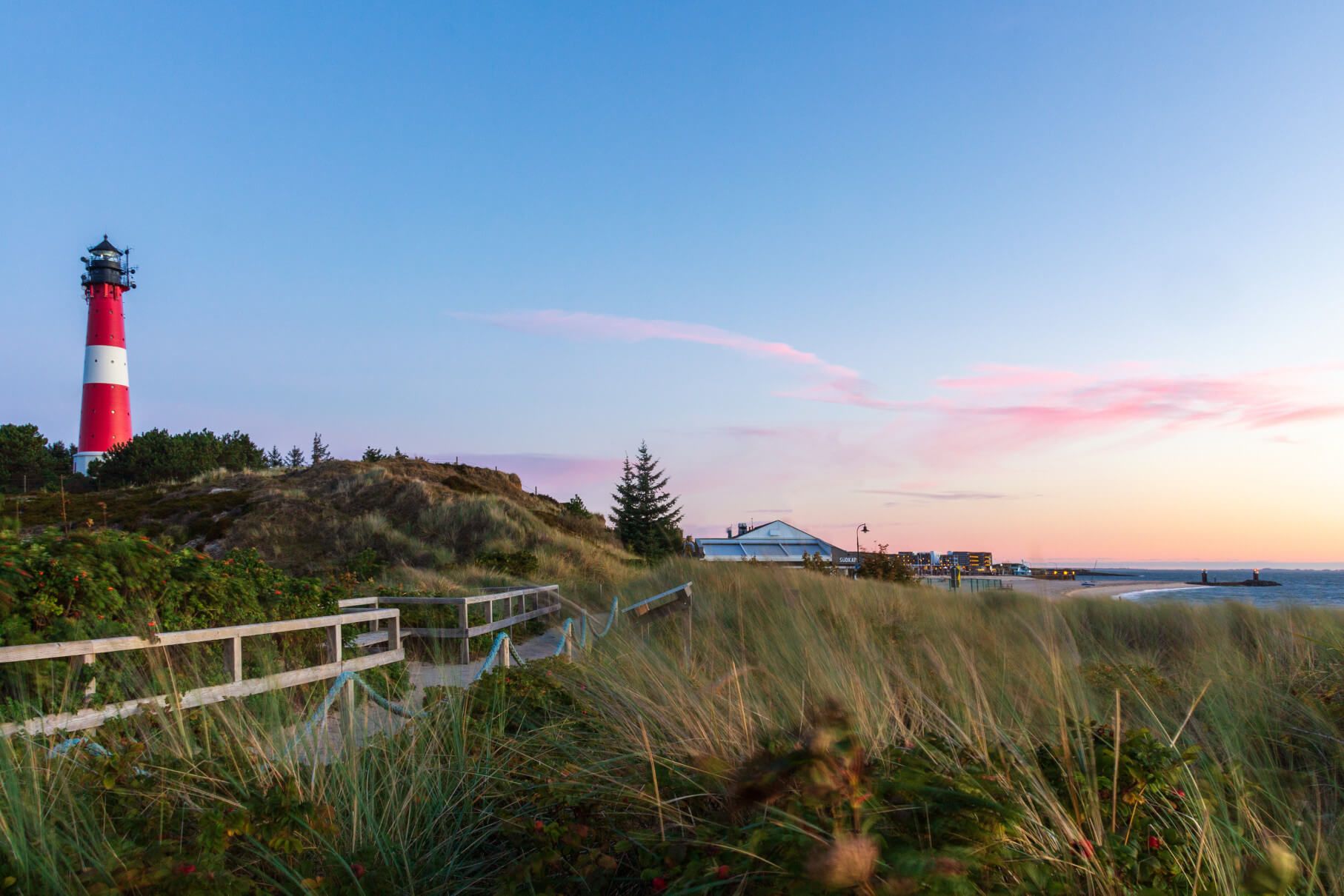 Ein roter Leuchtturm steht an der Küste, umgeben von Dünen und sanften Wellen unter einem pastelgerichteten Himmel.