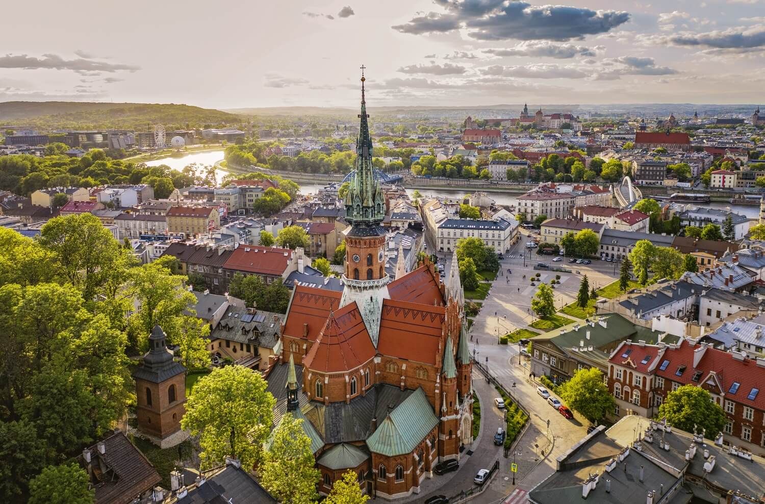 Blick auf eine Stadt mit einer markanten Kirche, Fluss, vielen Bäumen und historischen Gebäuden bei Sonnenuntergang.