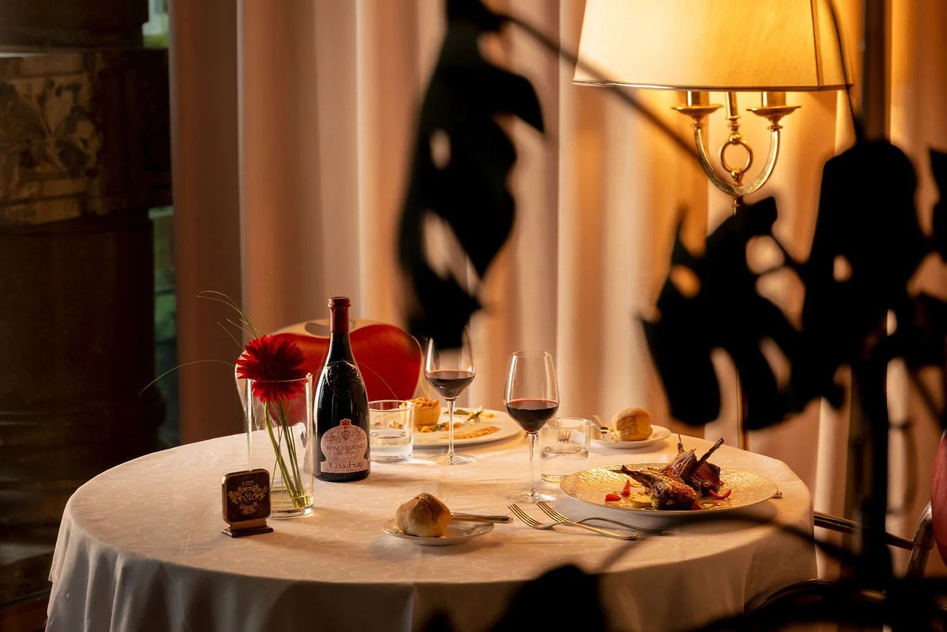 Elegant gedeckter Tisch mit Wein, köstlichem Essen und Blumen, beleuchtet von einer stilvollen Lampe.