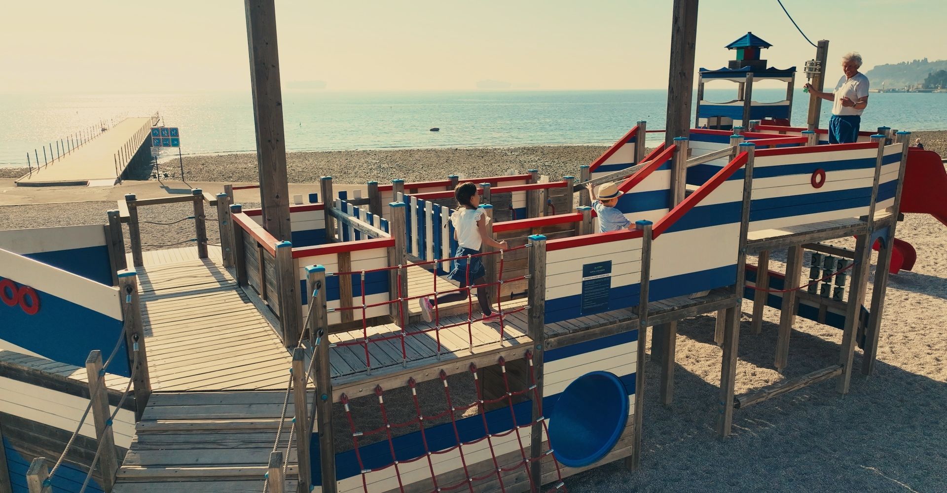 Ein Spielplatz im maritimen Stil am Strand, mit Kindern, die spielen und der Blick auf das ruhige Wasser.