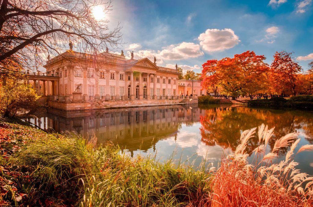 Ein prächtiges Gebäude spiegelt sich im ruhigen Wasser, umgeben von buntem Herbstlaub und strahlendem Himmel.