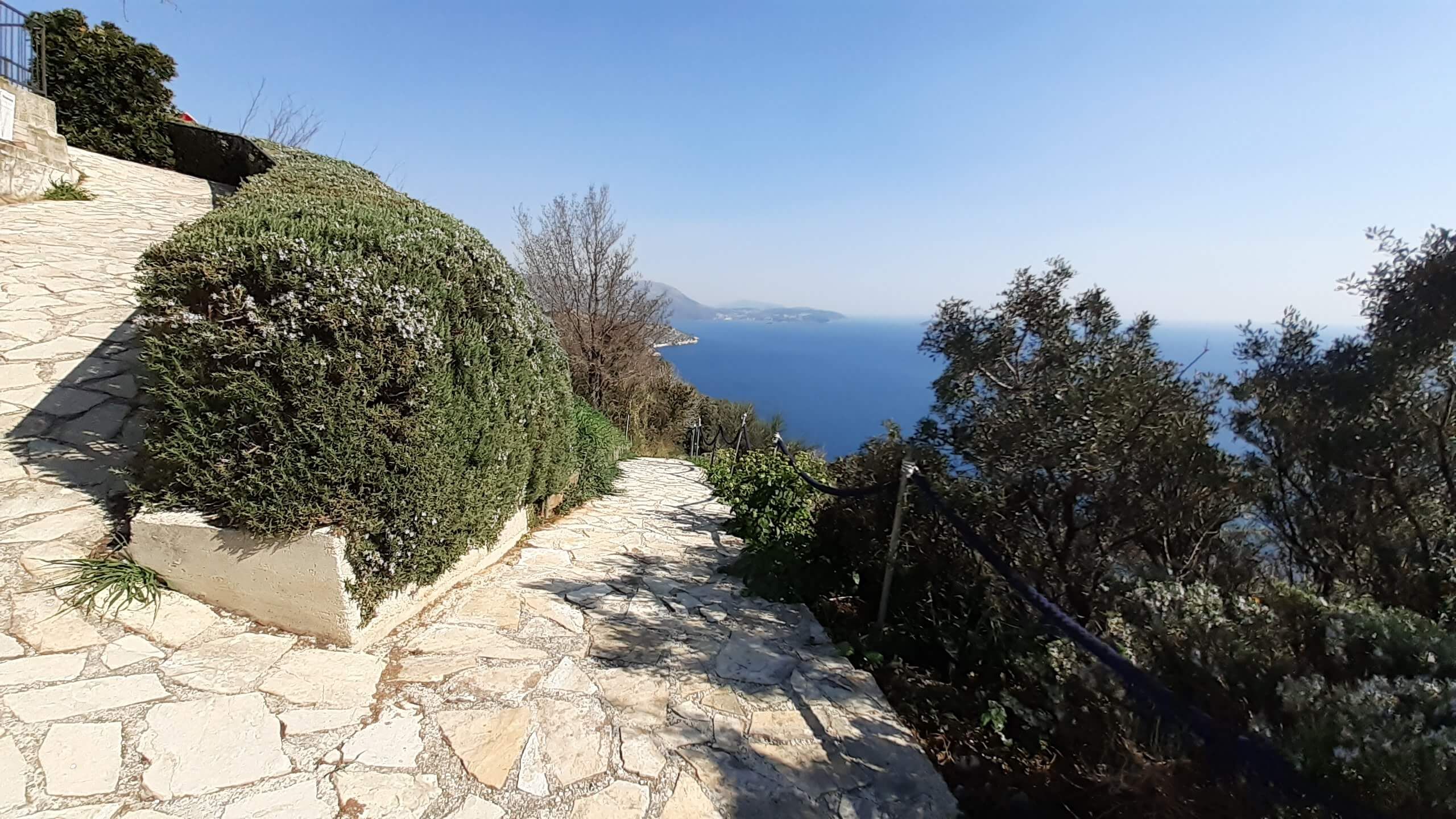 Ein malerischer Weg führt zum blauen Meer, umgeben von strauchigen Pflanzen und klarem Himmel.