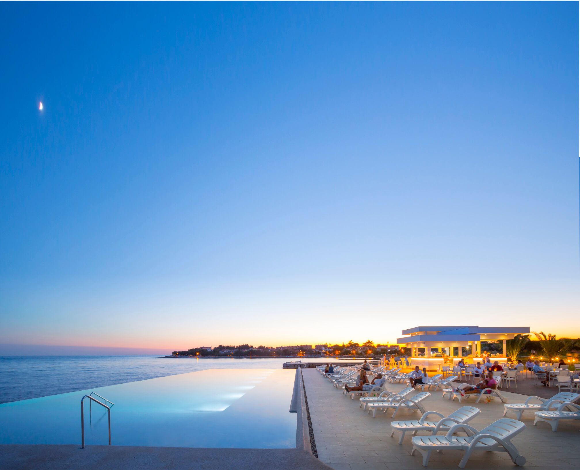 Ein ruhiger Abend am Meer mit einem Infinity-Pool, Liegen und einer Bar im Hintergrund unter klarem Himmel.