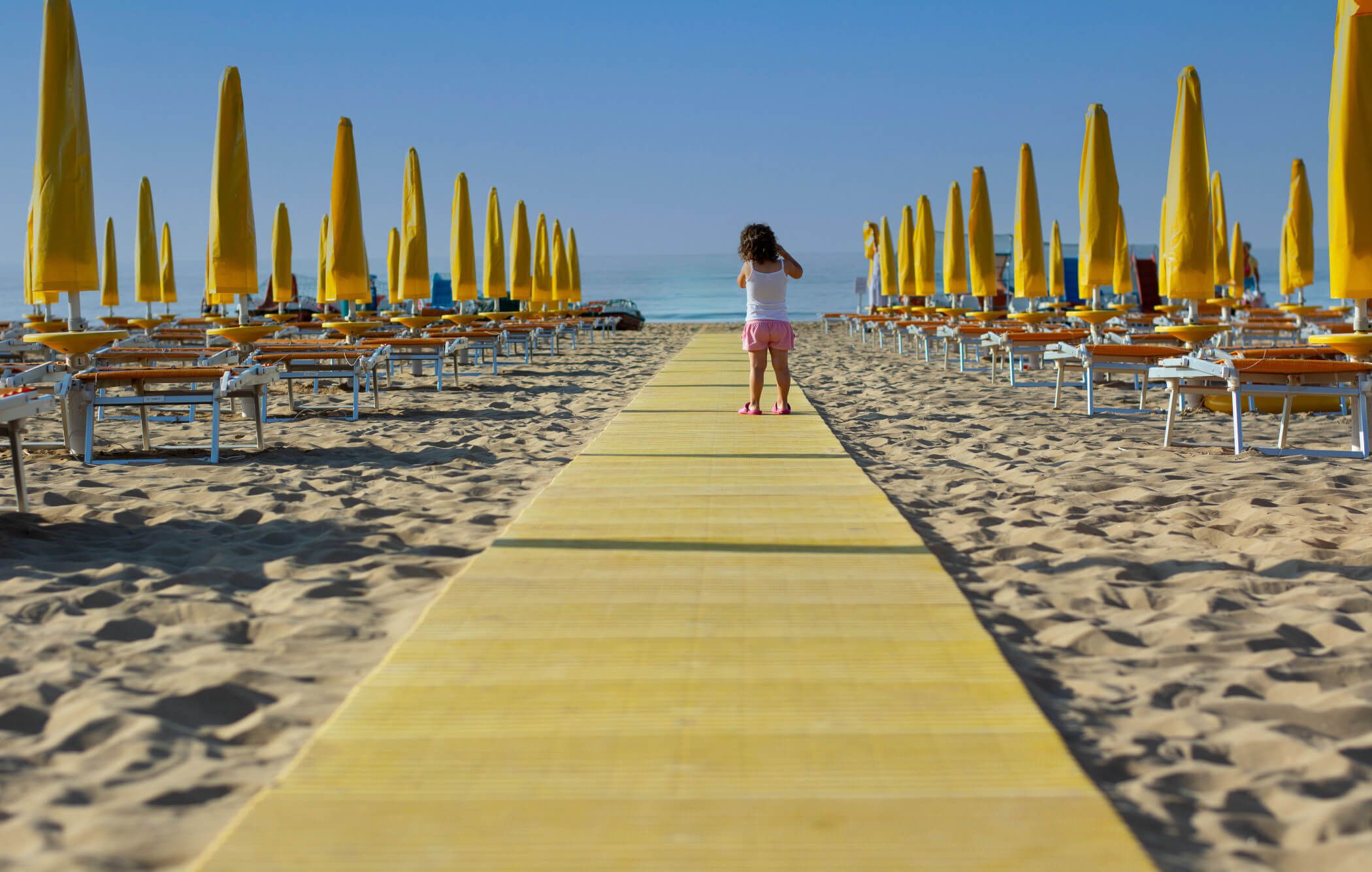 Ein Mädchen geht auf einem gelben Weg zwischen Liegen und Sonnenschirmen am Strand entlang, Meer im Hintergrund.