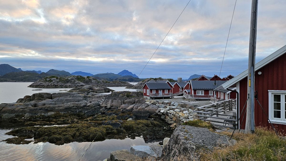 Rote Holzhütten am Wasser, umgeben von felsiger Küste und Bergen, unter einem bewölkten Himmel. Ruhe und Natur pur.