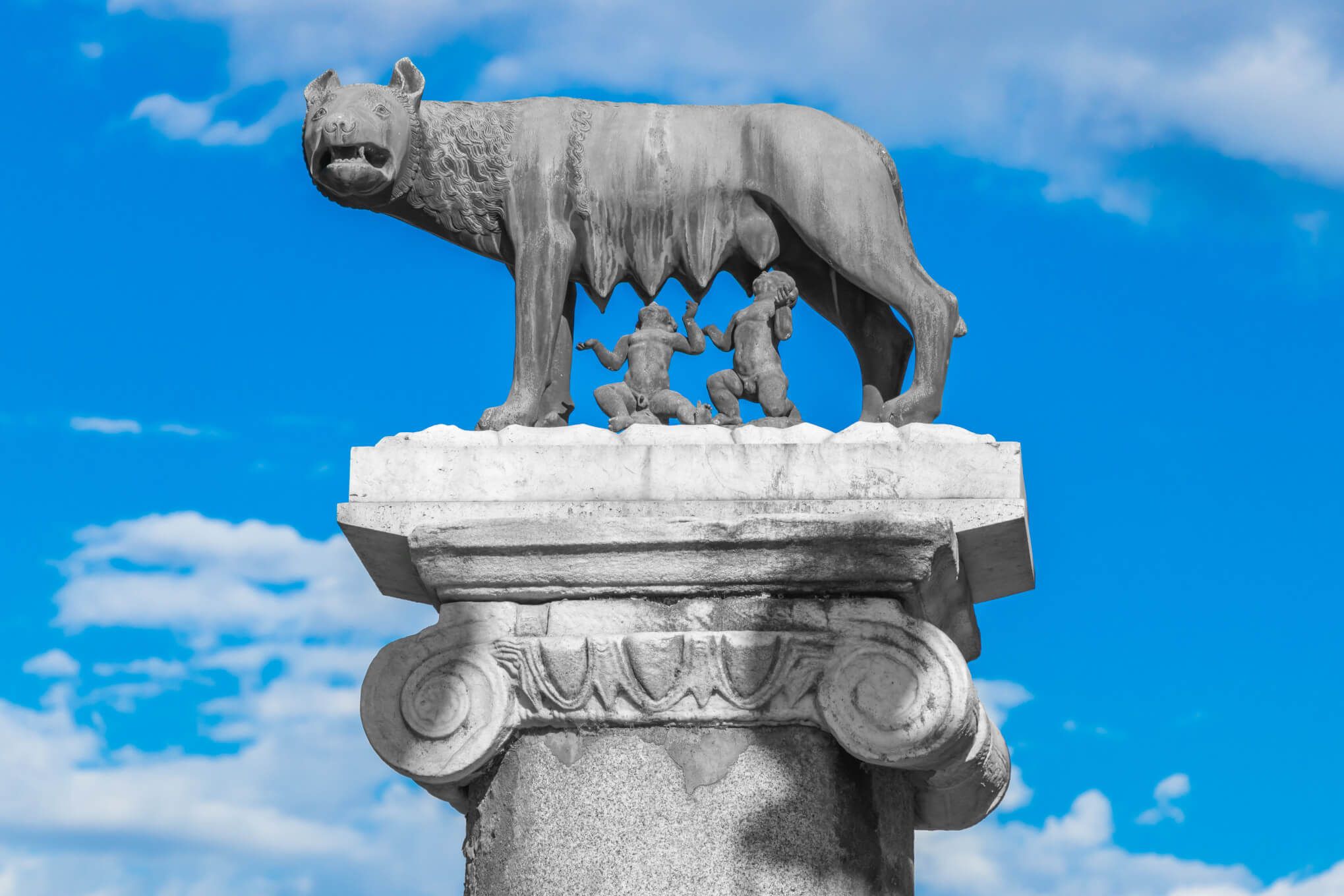 Eine Statue einer Wölfin mit zwei Säuglingen, die mythische Gründung Roms symbolisieren. Blauer Himmel im Hintergrund.