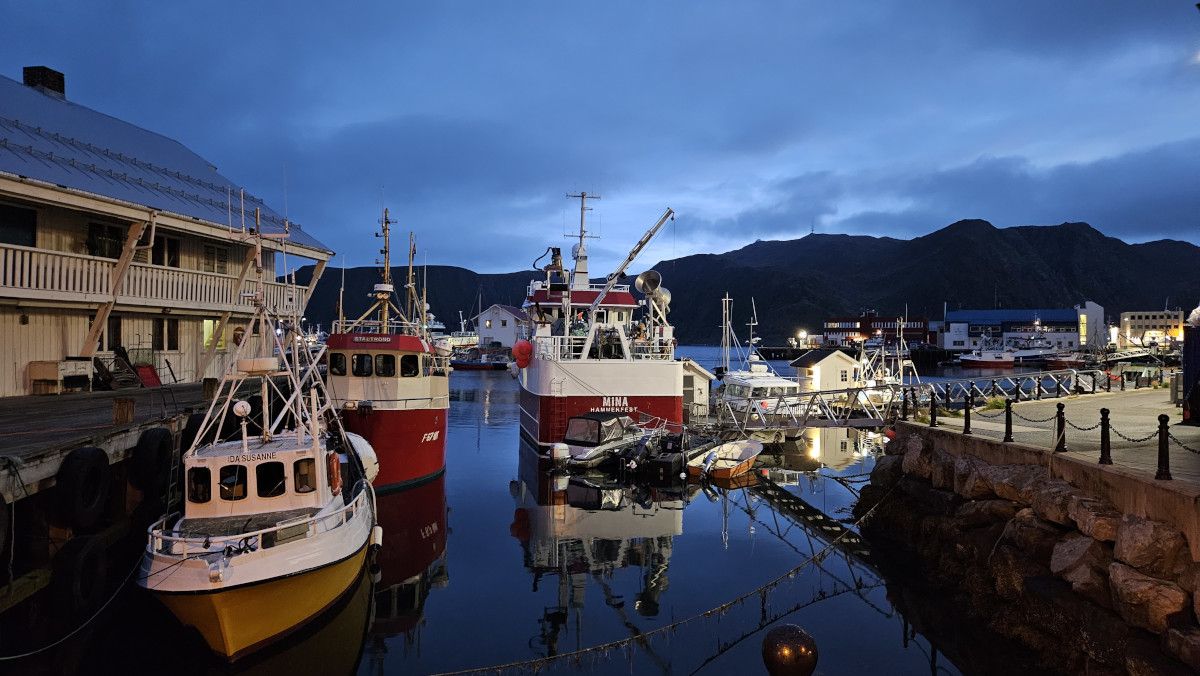 Ein Hafen bei Dämmerung mit Booten, reflektierendem Wasser und Bergen im Hintergrund. Ruhe und Idylle.