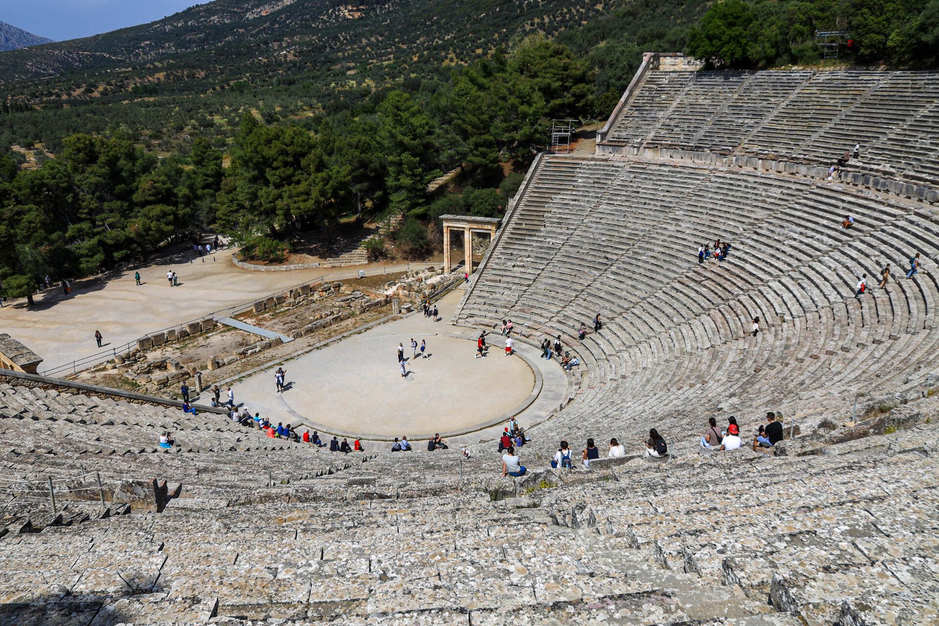 Ein antikes Theater in einer bergigen Landschaft, mit Besuchern auf den Sitzreihen und im unteren Bereich.