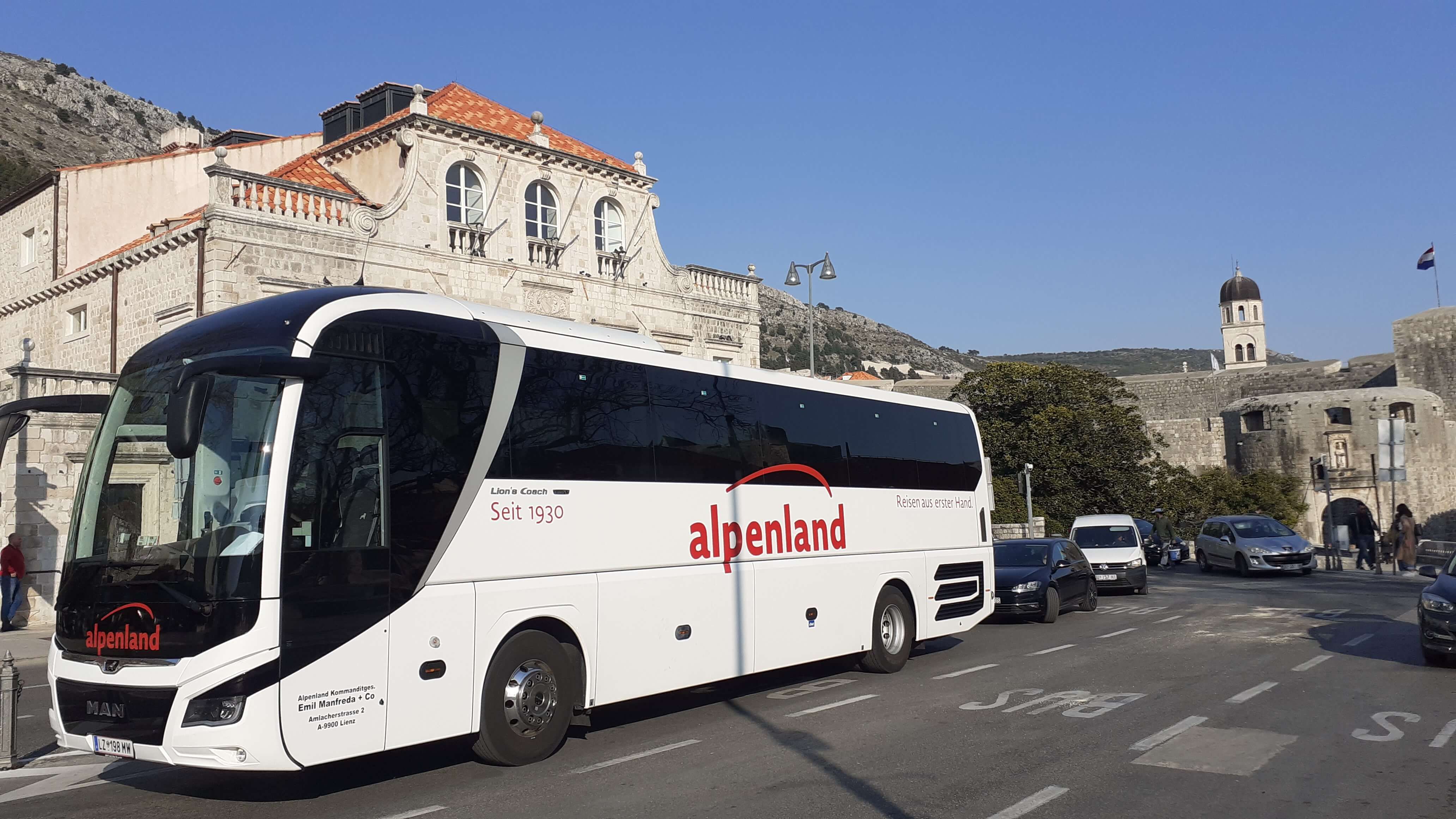Ein moderner Reisebus von Alpenland steht auf einer Straße vor historischen Gebäuden und Bergen im Hintergrund.