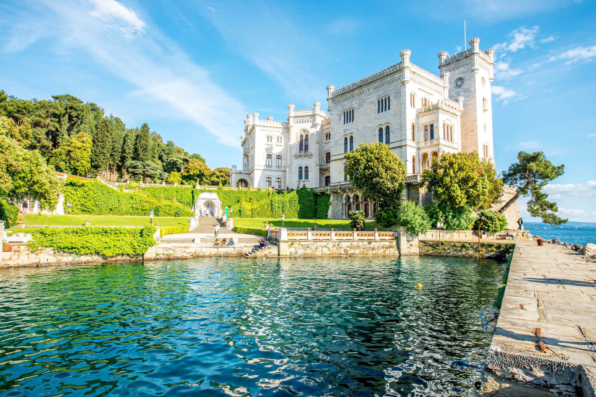 Das beeindruckendes Schloss Miramare in Triest, umgeben von grünen Bäumen, am Wasser mit klarem, blauem Himmel im Hintergrund.