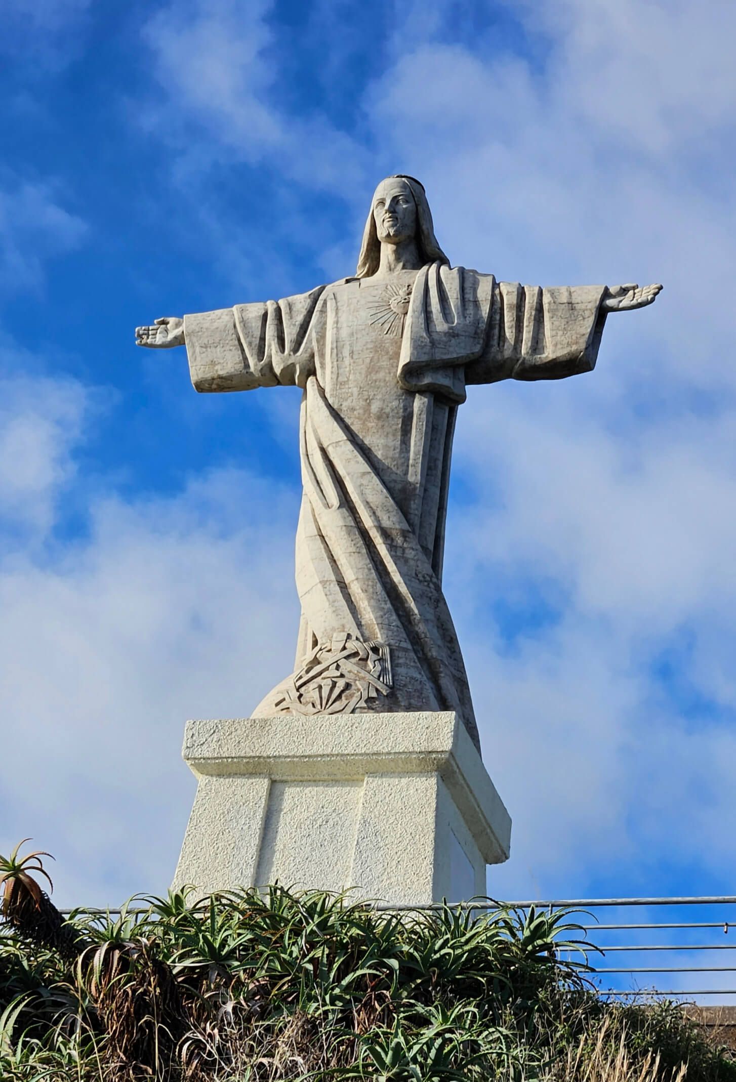 Eine majestätische Statue von Jesus Christus mit ausgebreiteten Armen vor blauem Himmel und Wolken.