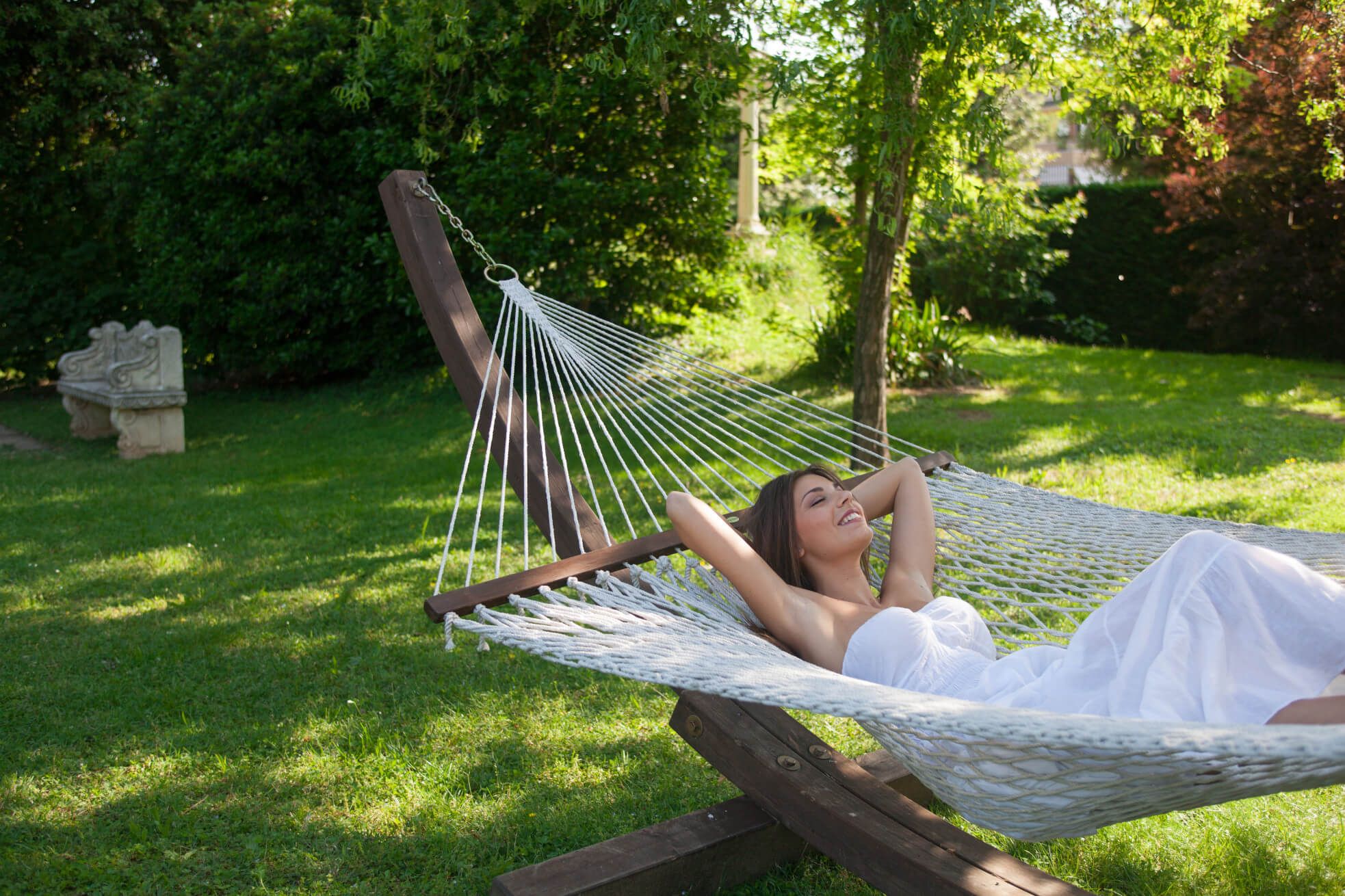 Eine Frau in einem weißen Kleid entspannt in einer Hängematte im grünen Garten und genießt die Sonne.