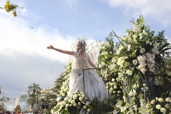 Eine elegante Frau in einem blumigen Kostüm posiert auf einem festlichen Wagen während eines Umzugs.