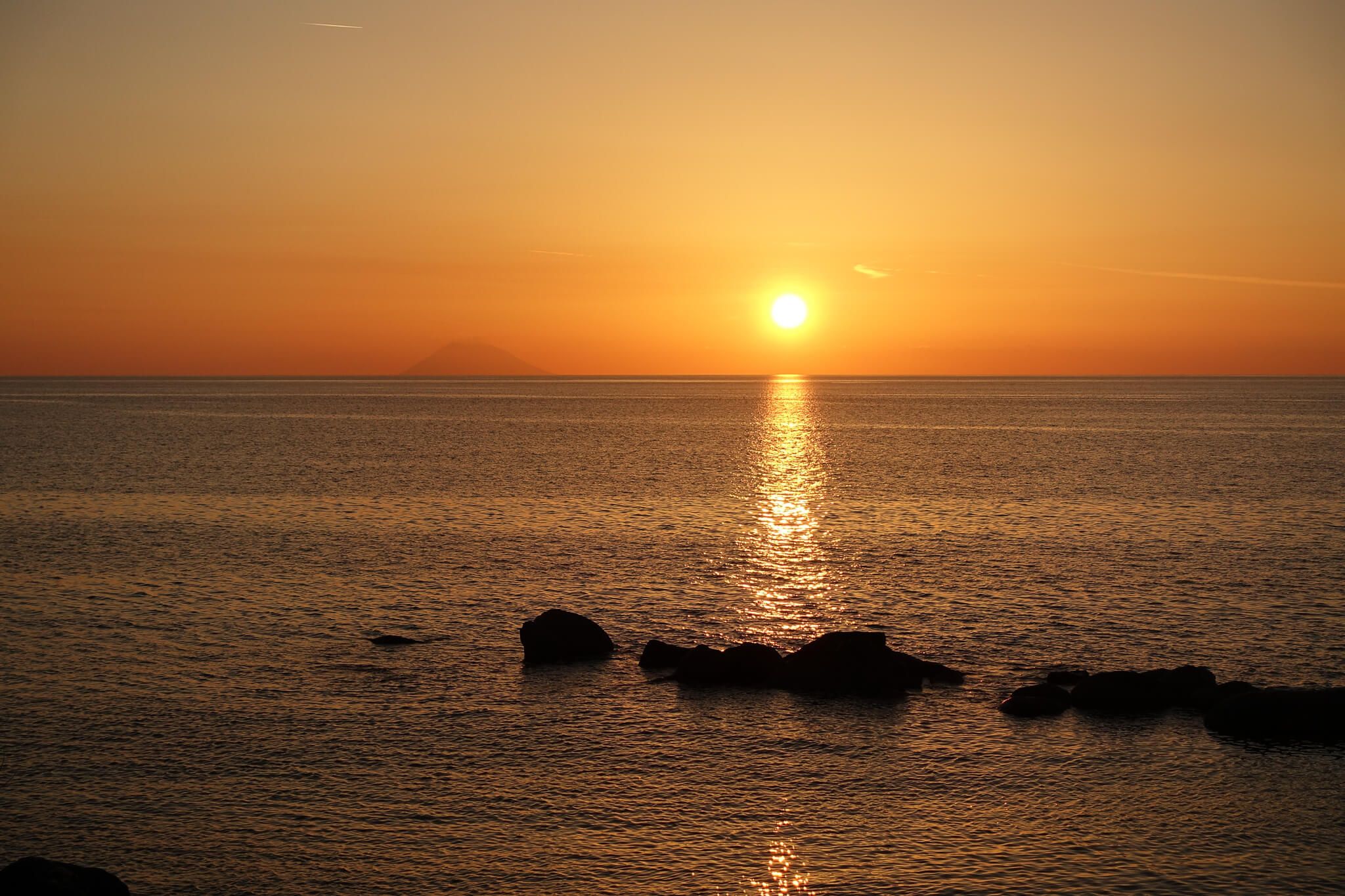 Sonnenuntergang über ruhigem Wasser, goldenes Licht spiegelt sich auf der Oberfläche, im Hintergrund eine Bergsilhouette.