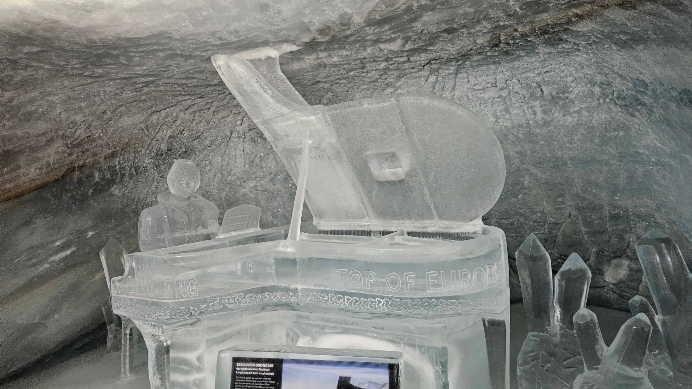 Eisskulptur eines Pianos mit einer Figur, umgeben von kristallartigen Eisformen, in einer Grotte.
